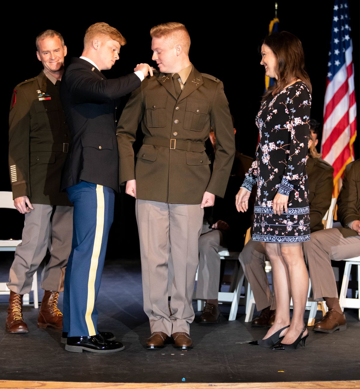 A military commissioning ceremony on a stage. A man in a dark blue dress uniform pins rank insignia onto the shoulder of a man in a tan and olive green Army Green Service Uniform. A woman in a floral dress stands to the right, smiling at the new officer, while an American flag is visible in the background.