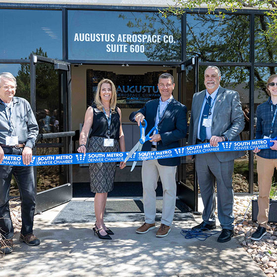 Ribbon cutting for Augustus Aerospace CO. with large blue scissors, ribbon that says " South Metro Denver Chamber" and owners of the business.