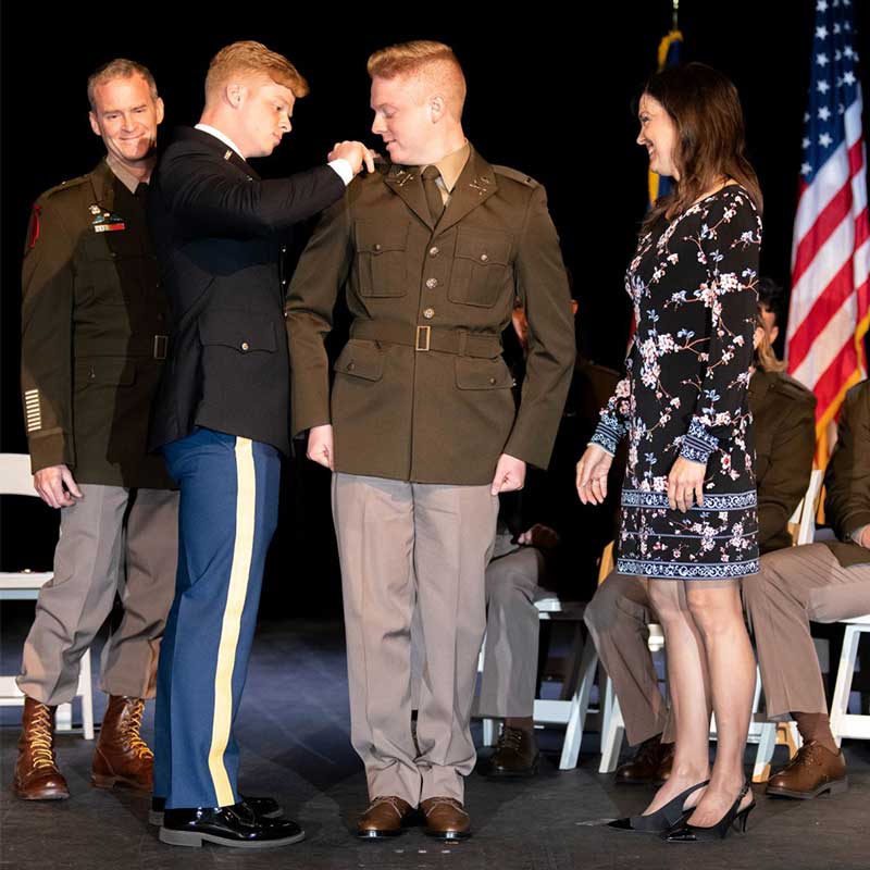 Celebration of a military commissioning ceremony on a stage. A man in a dark blue dress uniform pins rank insignia onto the shoulder of a man in a tan and olive green Army Green Service Uniform. A woman in a floral dress stands to the right, smiling at the new officer, while an American flag is visible in the background.